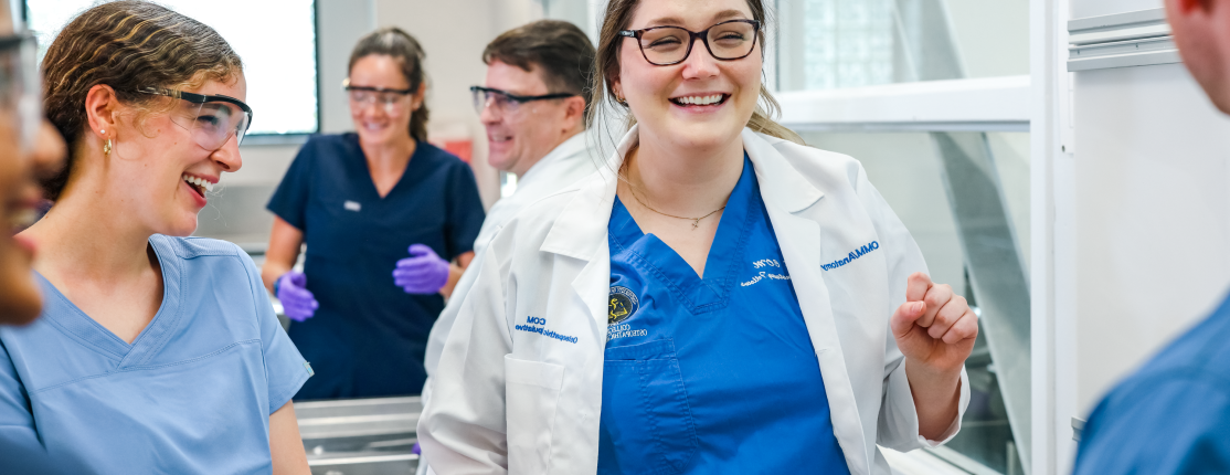 A student in a white coat talks with two other students in the anatomy lab